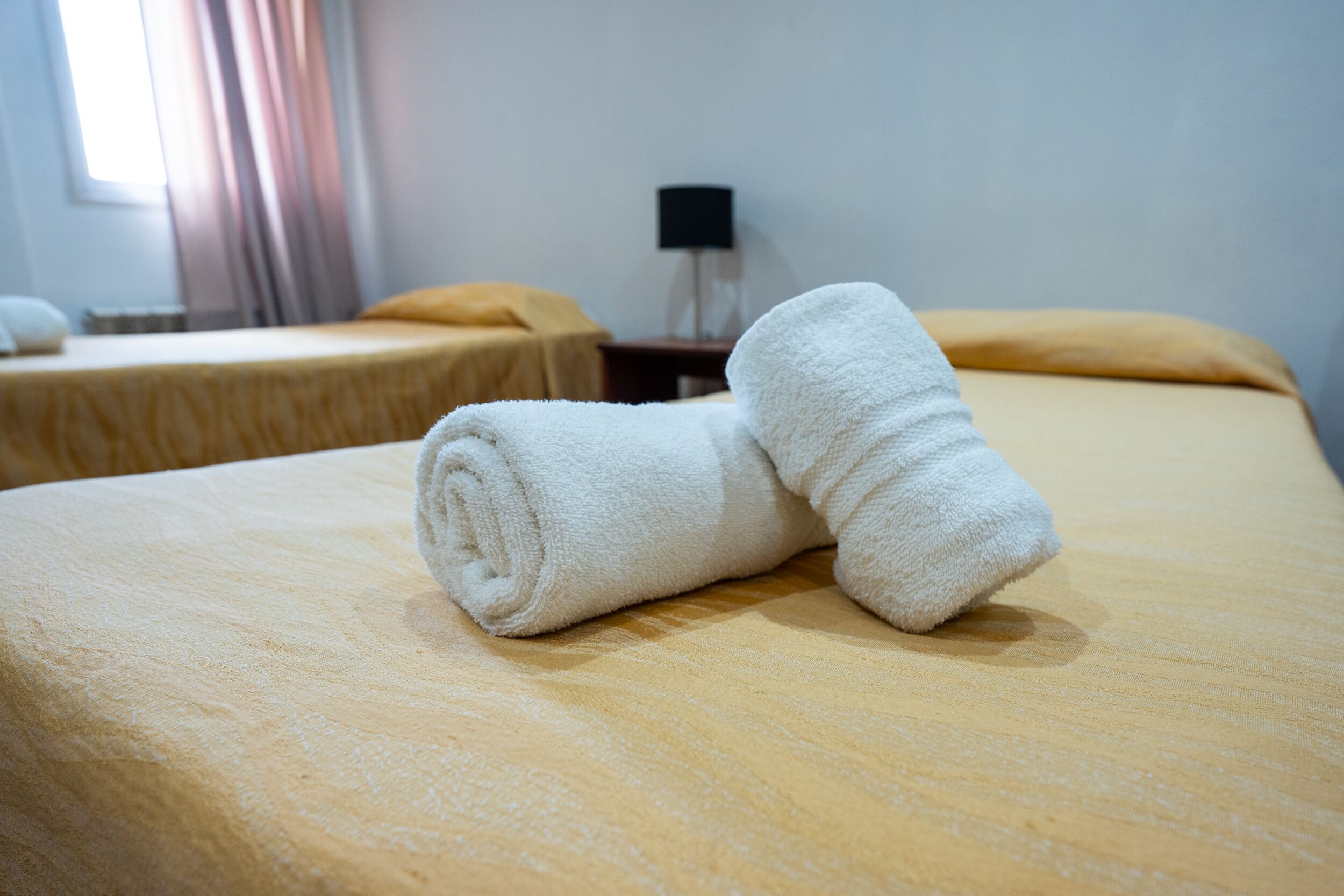 A hotel room with two beds, each with a yellow bedspread and a rolled white towel on one bed.