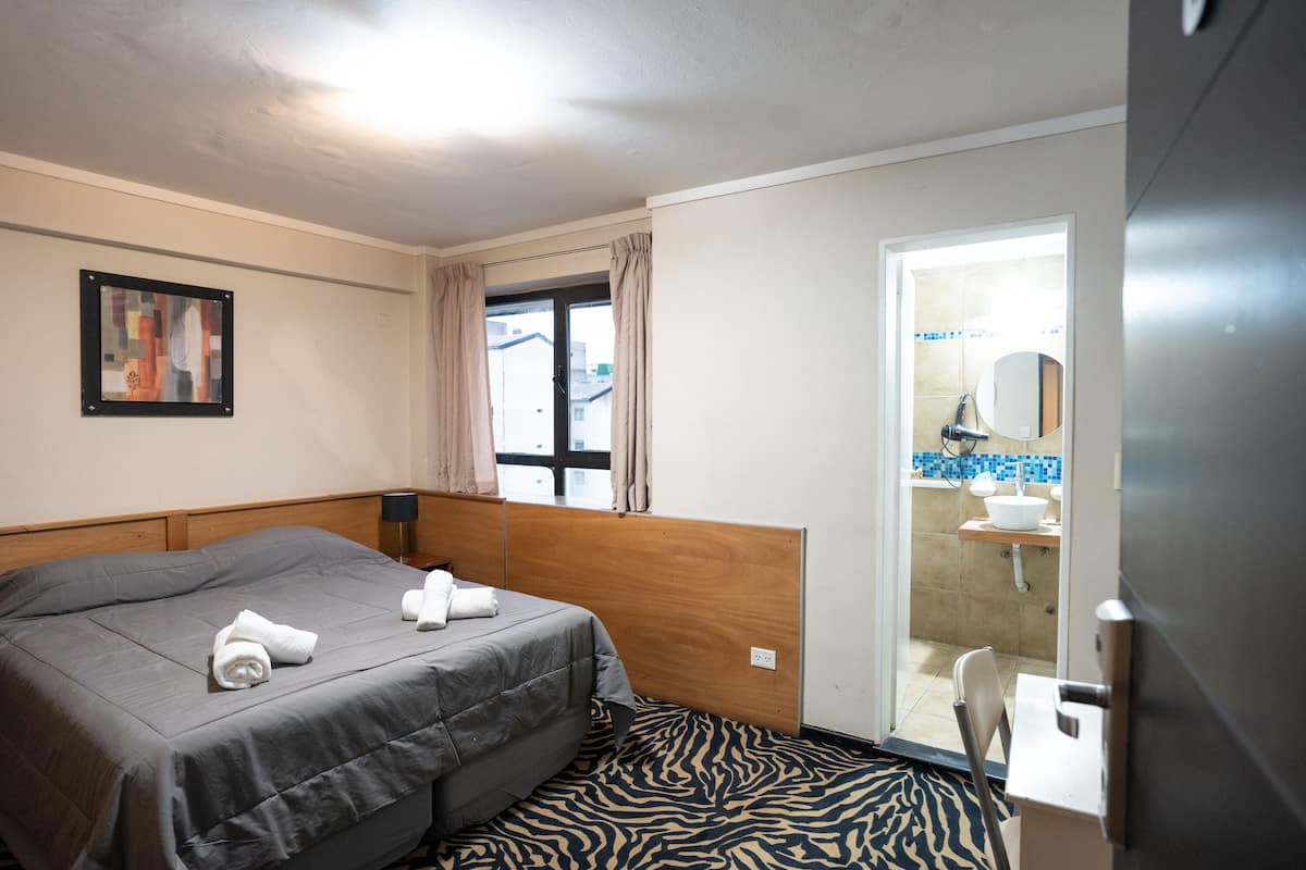 Habitación de hotel con cama, ventana con cortinas, baño visible a través de una puerta abierta y una alfombra con estampado de cebra.
