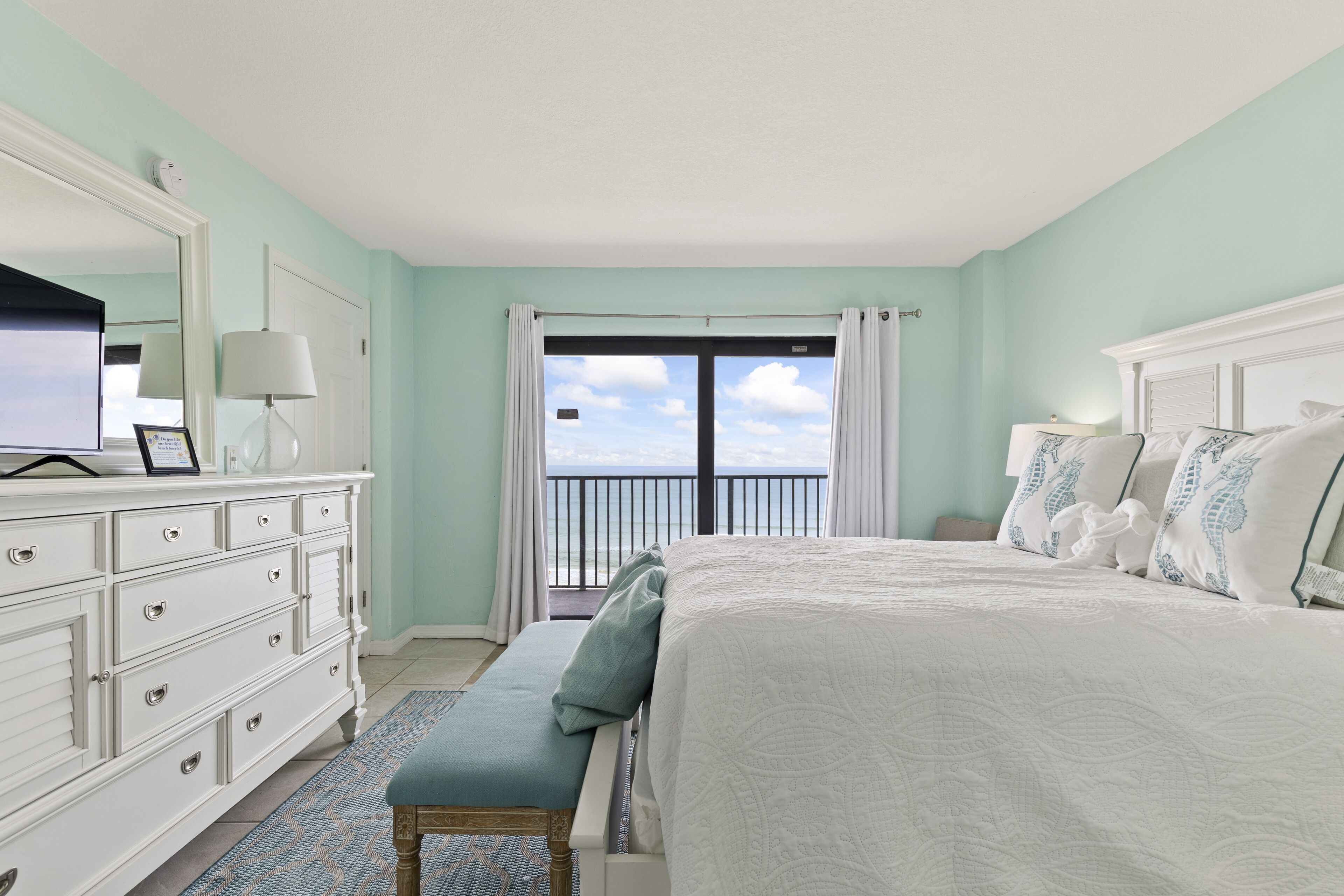 A bedroom with a large bed, a dresser, a bench, a TV, and a balcony with a view of the sky.