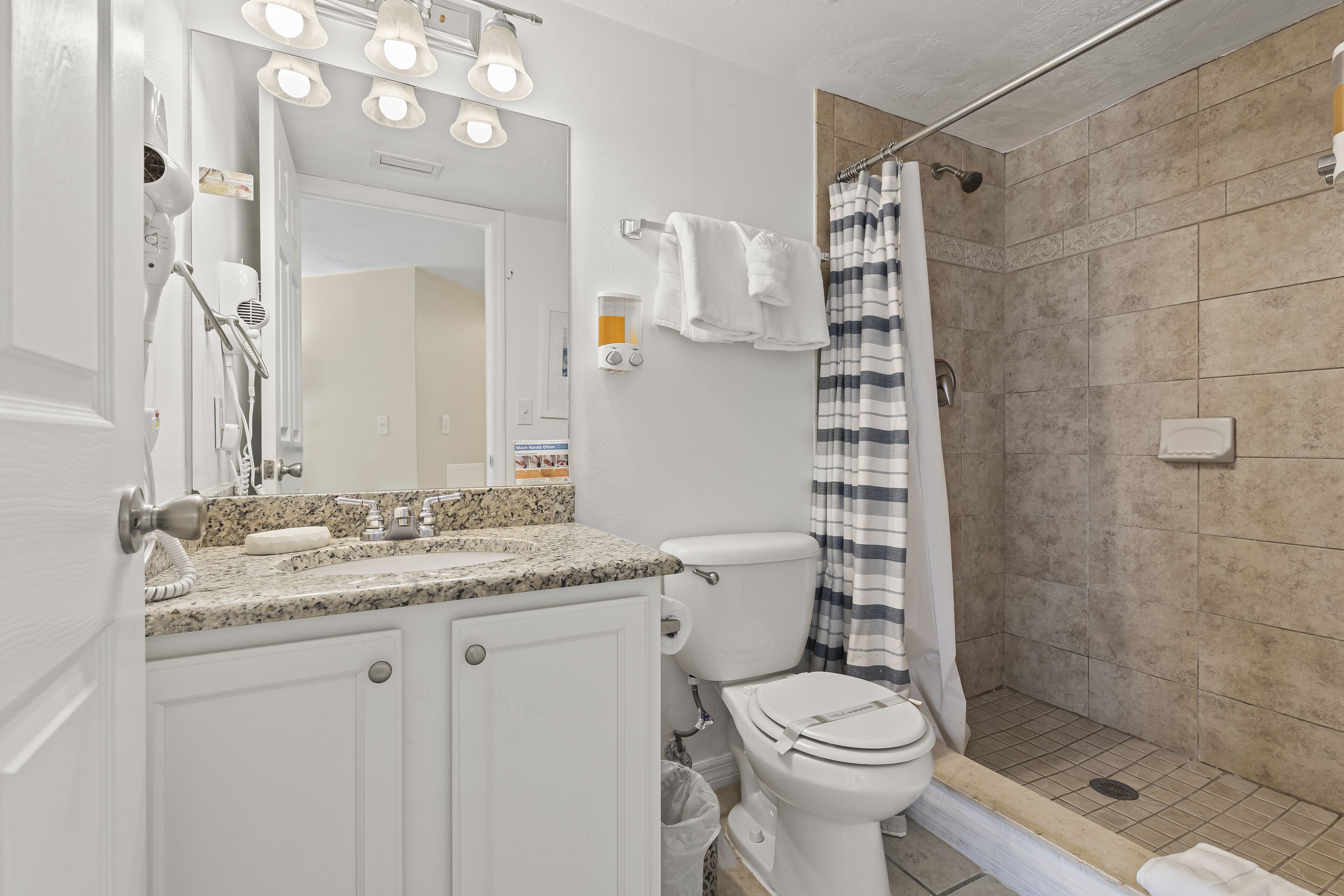 Une salle de bain avec un comptoir en granit, des toilettes, une douche avec un rideau à rayures et un miroir avec éclairage.