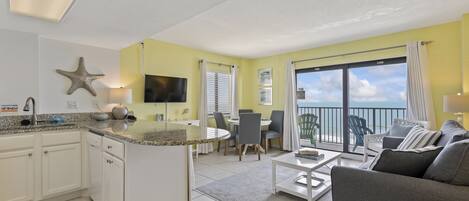 A modern kitchen with white cabinets, a granite countertop, and a starfish decoration. There is a dining area with a table and chairs, and a balcony with a view of the ocean.