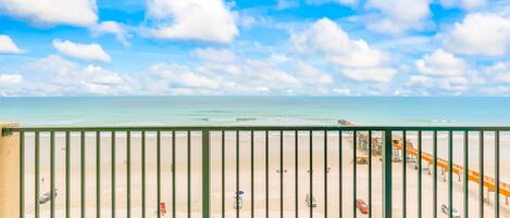 A beach view from a balcony with a railing, a sandy beach, and a clear sky.