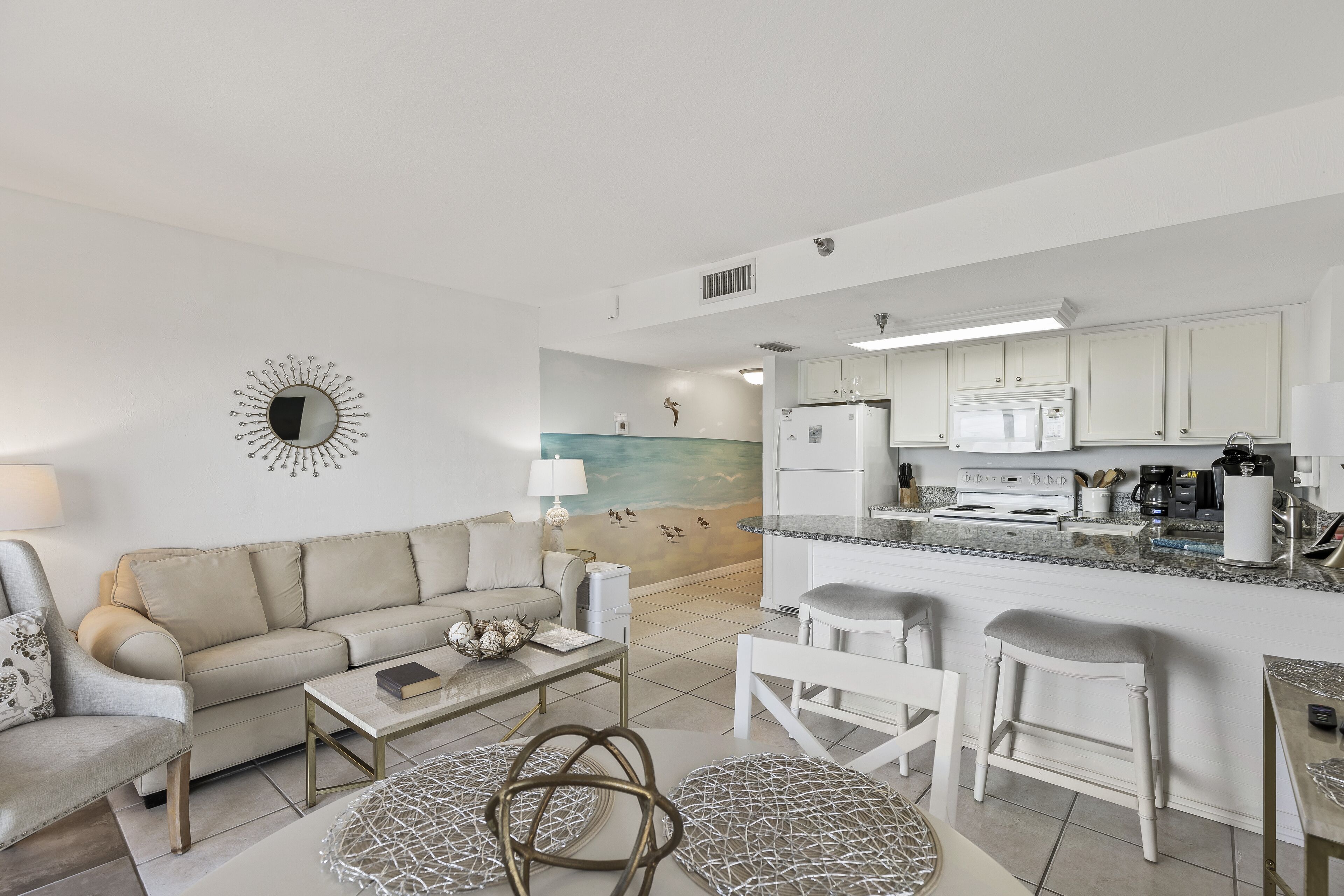 A modern living room with a sofa, coffee table, and a dining area with stools. A kitchen with white cabinets and appliances is visible in the background.