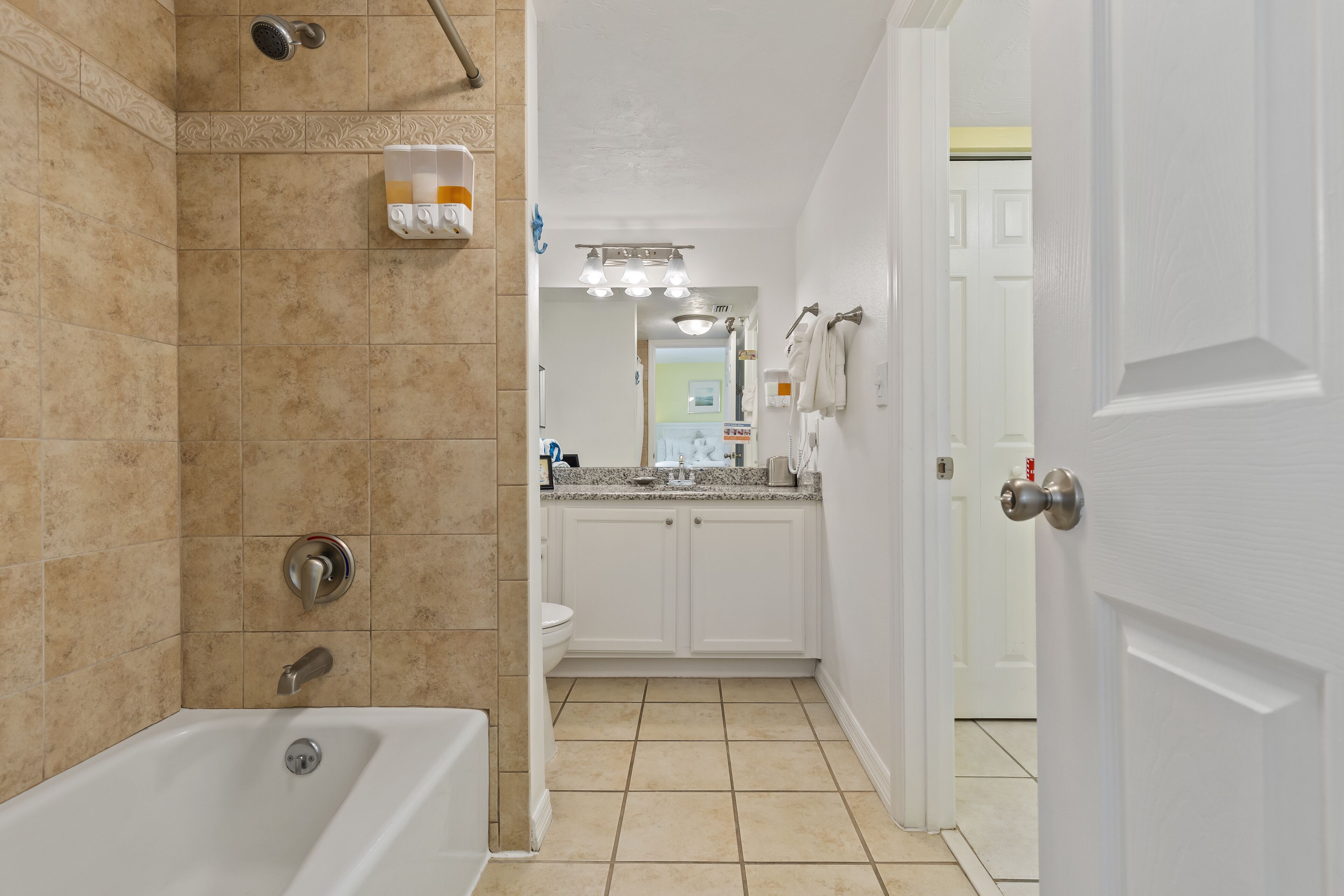 A bathroom with a bathtub, a shower, and a toilet. There is a sink with a countertop and a mirror above it. The walls are tiled, and there is a door leading to another room.