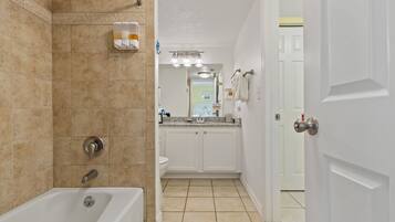 A bathroom with a bathtub, a shower, and a toilet. There is a sink with a countertop and a mirror above it. The walls are tiled, and there is a door leading to another room.