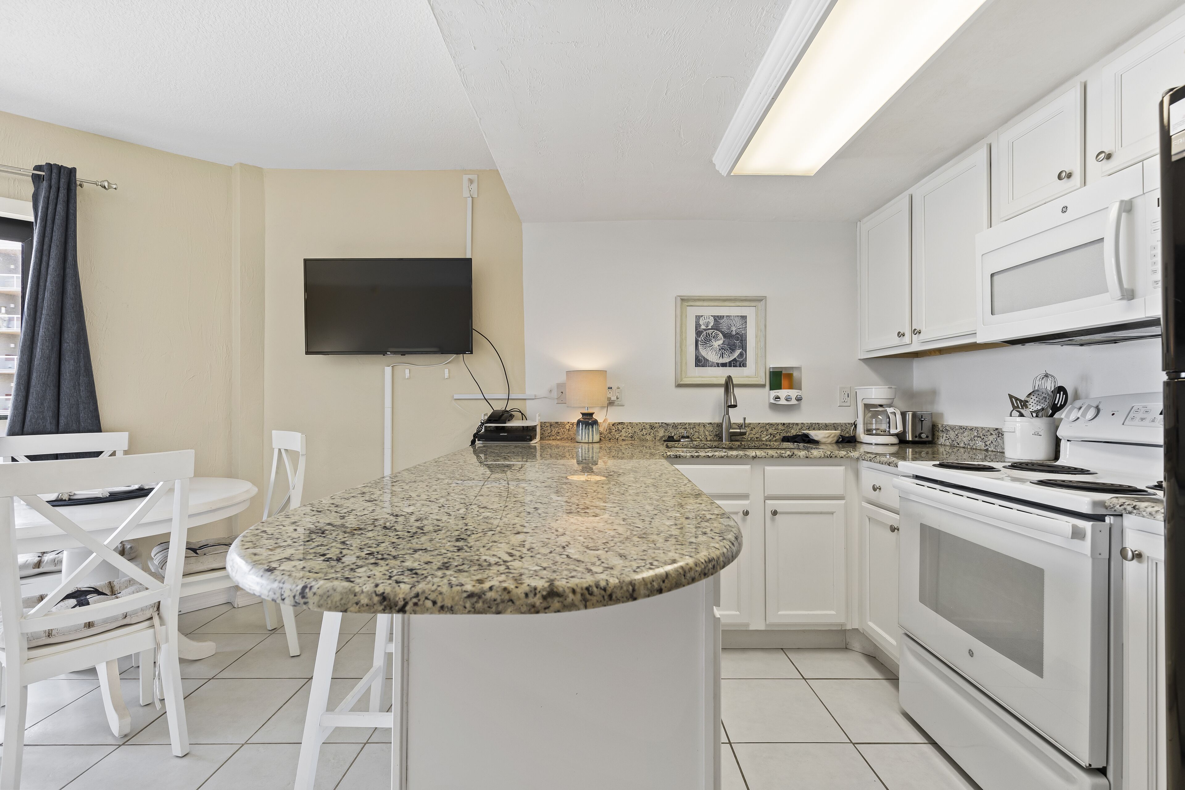 A modern kitchen with white cabinets, a granite countertop, a built-in oven, and a flat-screen TV.