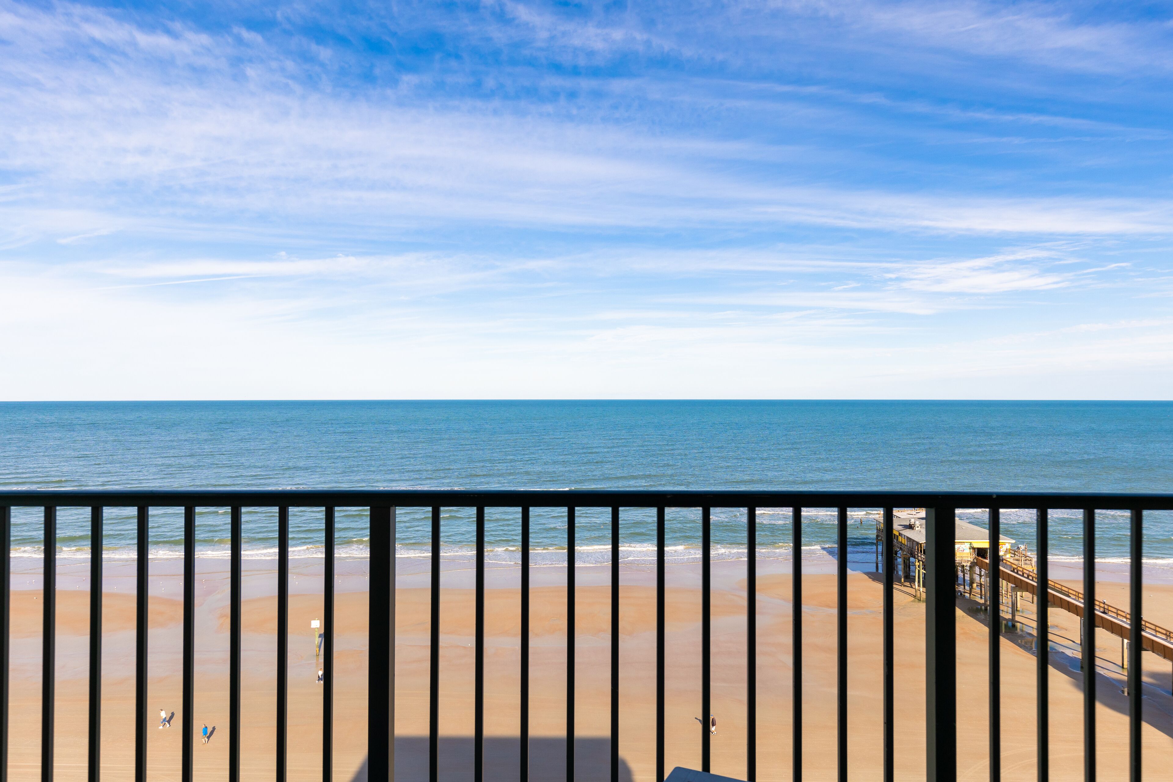 Vista de una playa desde un balcón con barandilla, una playa de arena y un muelle a lo lejos.