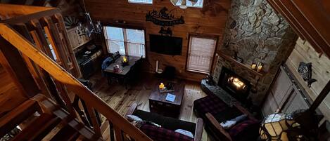 A cozy cabin interior with a wooden staircase, a stone fireplace, a dining area, and a ceiling fan.