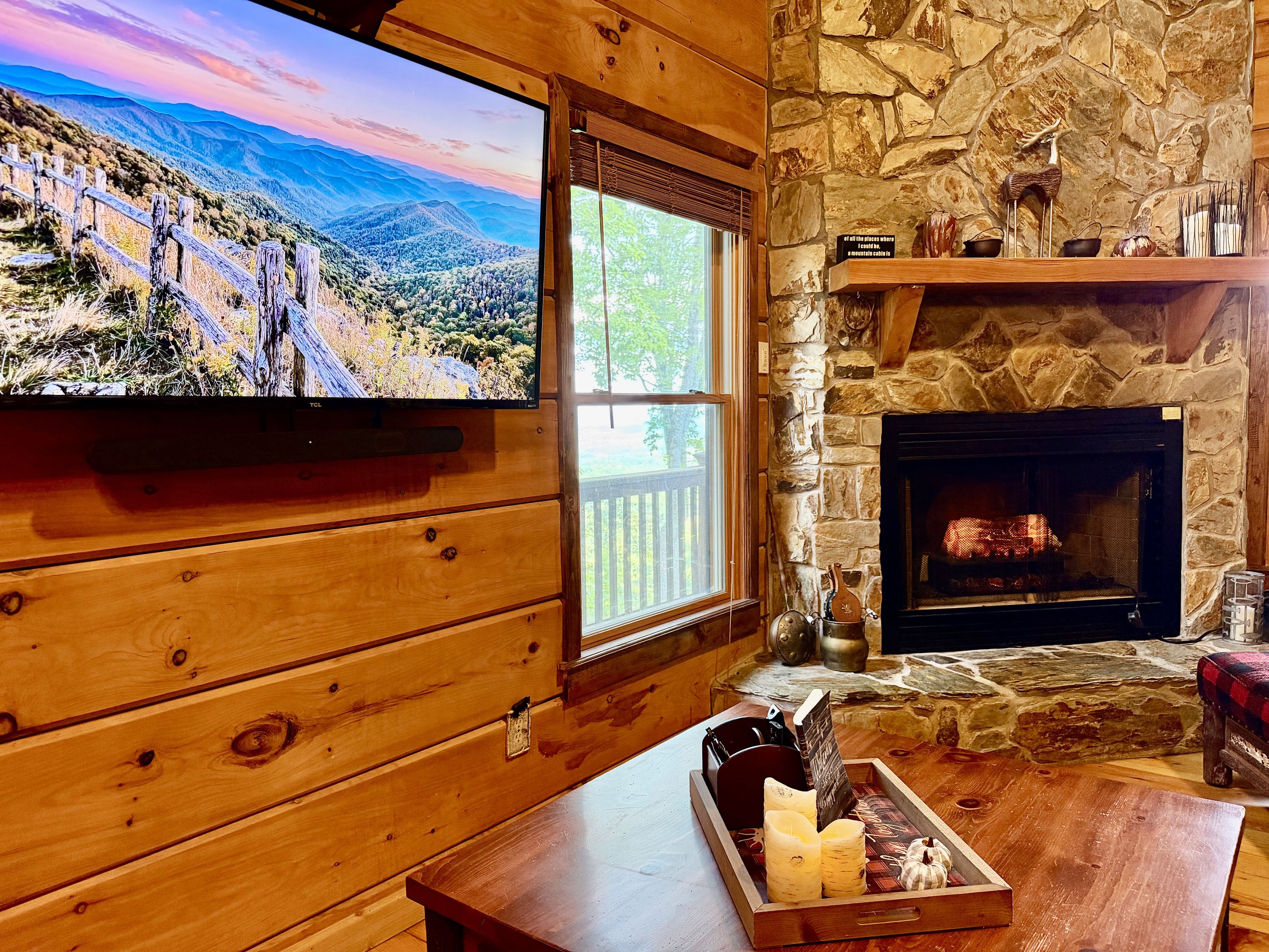 A cozy cabin interior with a stone fireplace, wooden walls, a television mounted on the wall, and a window overlooking a mountainous landscape.