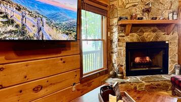 A cozy cabin interior with a stone fireplace, wooden walls, a television mounted on the wall, and a window overlooking a mountainous landscape.