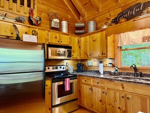 A kitchen with wooden cabinets, stainless steel appliances, and a window with a view of greenery. - JuBela's Cabin + Mountaintop Lake Views + Private Hot Tub + ASKA Trail Access (Blue Ridge)