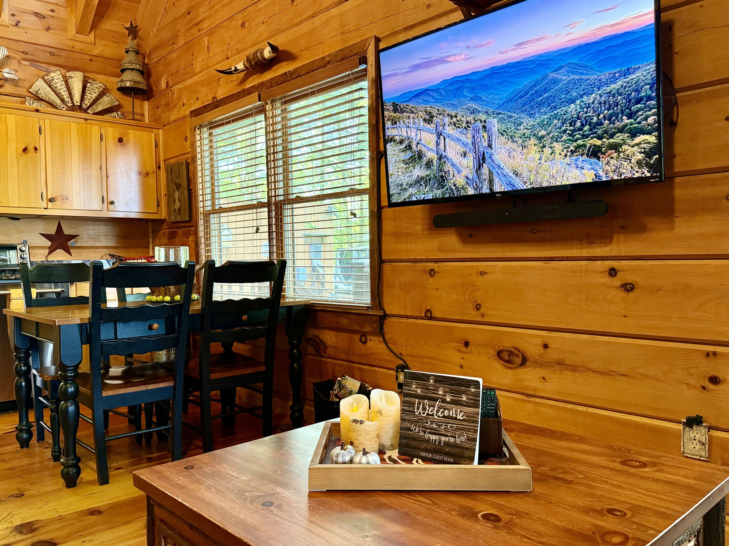 Ein Holzinterieur mit einem Fernseher, der eine Berglandschaft zeigt, einem Holztisch mit Kerze und einer Willkommensschild, sowie einer Sternen-Dekoration an der Wand.