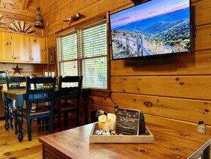A wooden interior with a television displaying a mountainous landscape, a wooden table with a candle and a welcome sign, and a star decoration on the wall. - JuBela's Cabin + Mountaintop Lake Views + Private Hot Tub + ASKA Trail Access (Blue Ridge)