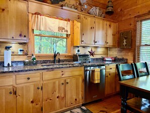 A wooden cabin kitchen with a granite countertop, stainless steel appliances, and a window with a view of trees.