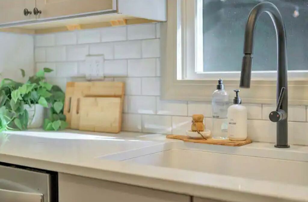 A kitchen with a white countertop, a wooden cutting board, a sink with a faucet, and a window above the sink.