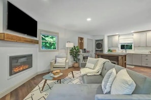 Cozy living room with a fireplace, sofa, and TV, featuring a kitchen area with white cabinets and countertop.