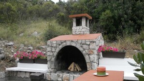 A stone-built outdoor fireplace with a chimney, surrounded by a garden with pink flowers, and a table with a candle.