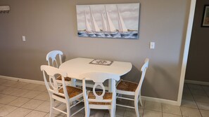 A dining area with a white table and chairs, a painting of sailboats on the wall, and a tiled floor. - Beach Front~2 BDRM Condo, 2 BA, Pool, 4 Bikes & VIEWS! One Level!  #1F (Hilton Head Island)