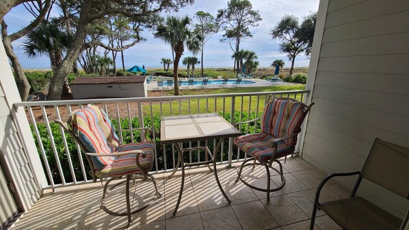 A balcony with two striped chairs, a small table, and a view of a swimming pool and beach. - Beach Front~2 BDRM Condo, 2 BA, Pool, 4 Bikes & VIEWS! One Level!  #1F (Hilton Head Island)