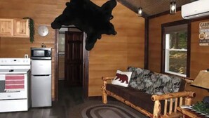 A cabin interior with a kitchen area, a wooden dining table, and a sofa with a bear pillow.