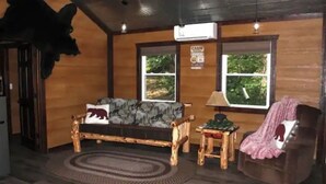 A cabin interior with a wooden sofa, a chair, a small table, and a window with a view of greenery.