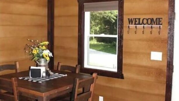 A wooden interior with a round dining table, chairs, a welcome sign, and a window with a view.