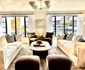 A modern living room with two sofas, a coffee table, and two armchairs. Large windows offer views of the outside.