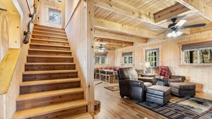 Un escalier en bois mène à une chambre à coucher située en mezzanine, dotée d’un ventilateur de plafond et offrant une vue sur l’extérieur.