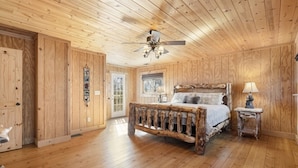 Une chambre à coucher aux murs lambrissés, avec un lit rustique, un ventilateur de plafond et une petite table de chevet surmontée d’une lampe.