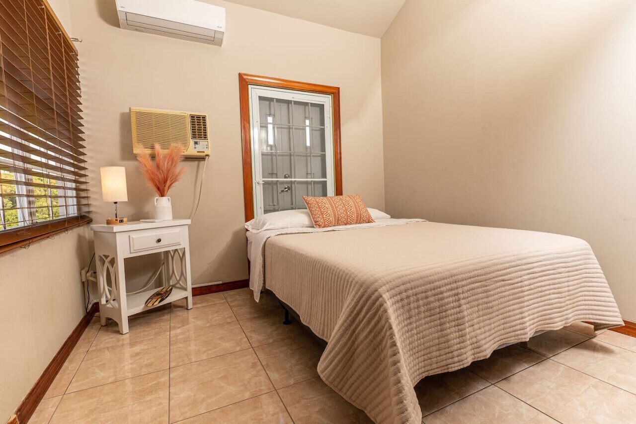 Camera da letto con un letto, un comodino, una lampada, un condizionatore d'aria, una finestra e uno specchio fissato alla parete.
