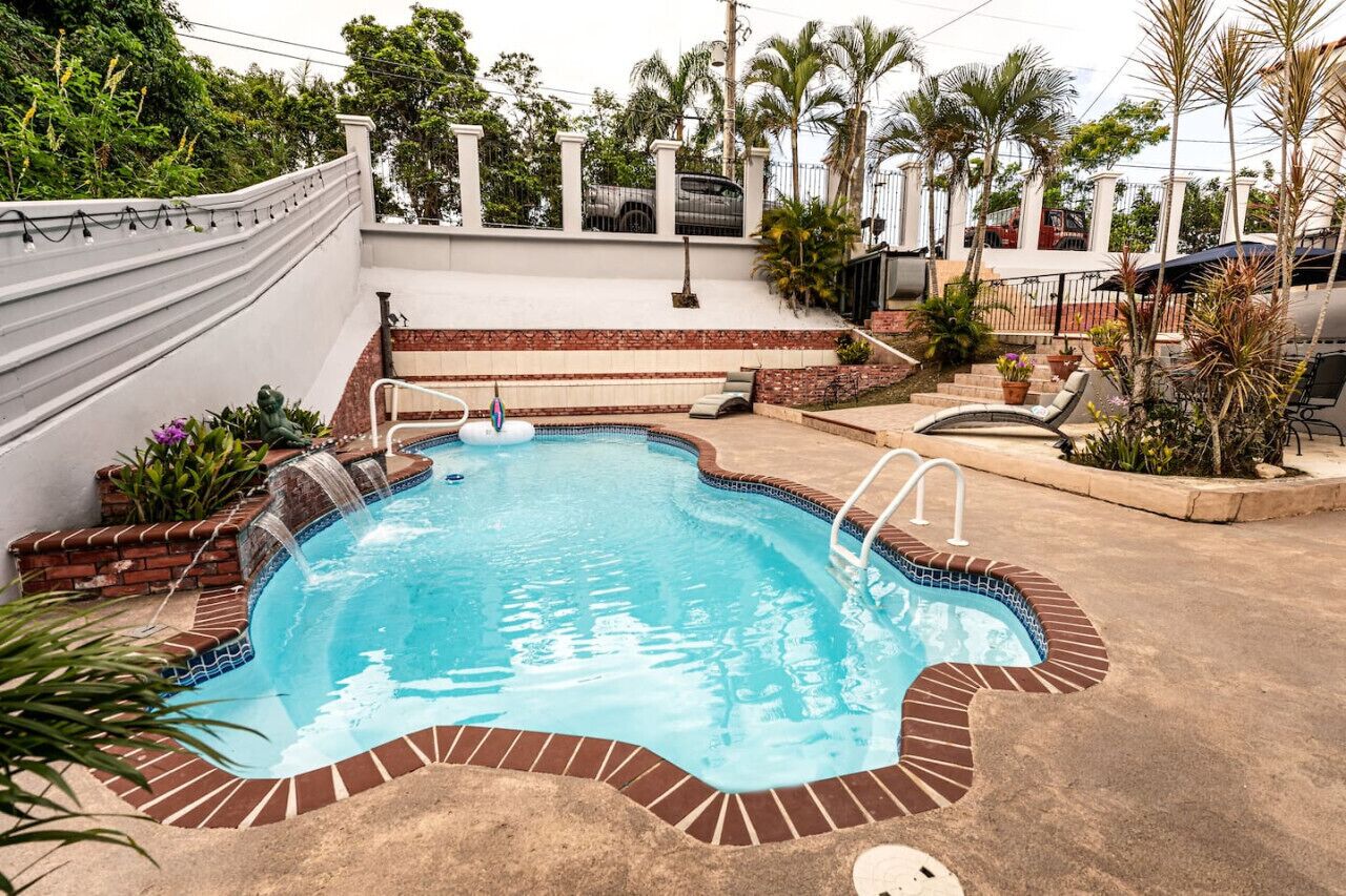 Una piscina con borde de ladrillo, una cascada y un patio circundante con plantas y mobiliario de exterior.