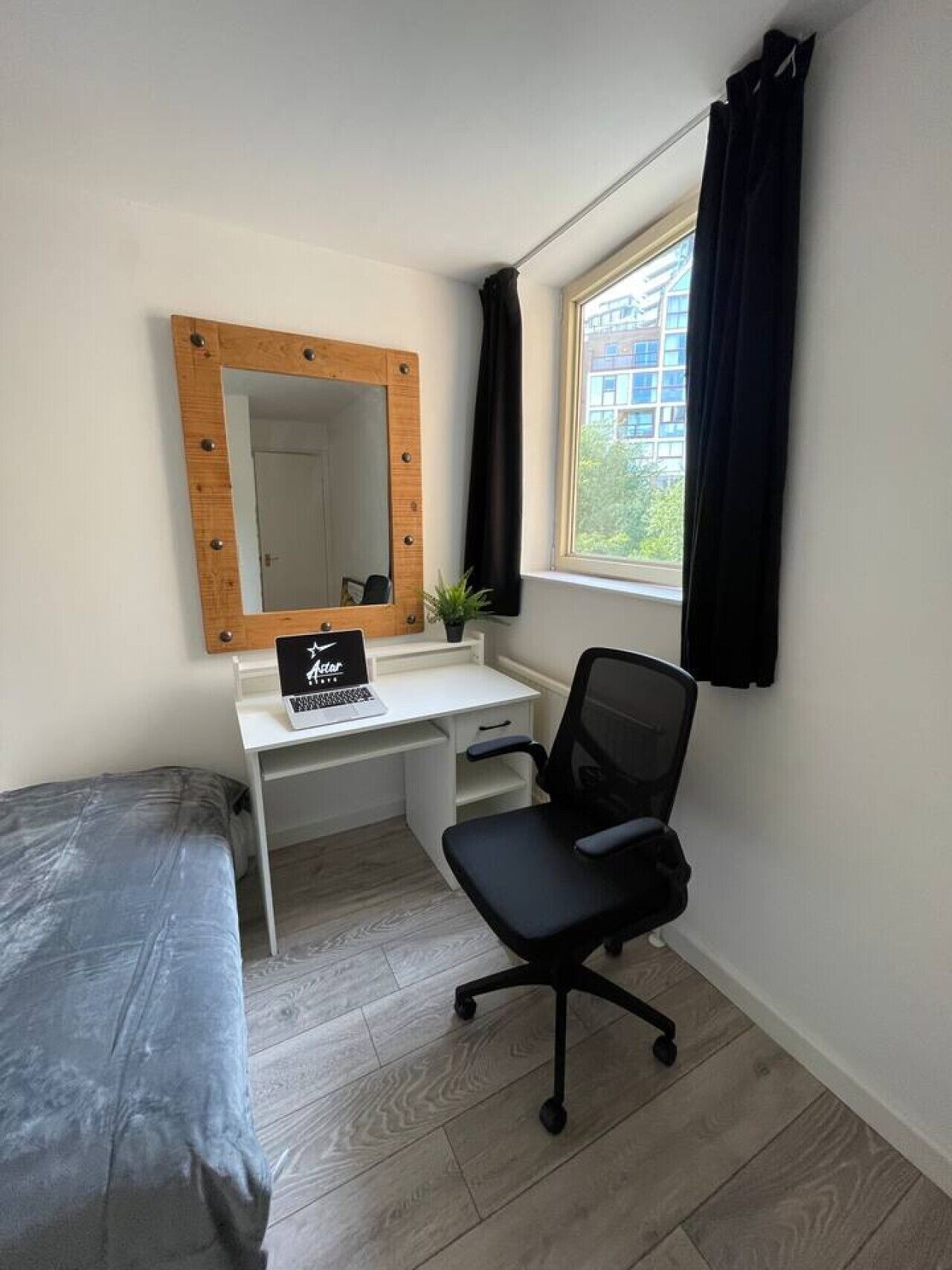 Un dormitorio con cama, un escritorio con una computadora portátil, una silla, un espejo y una ventana con cortinas.