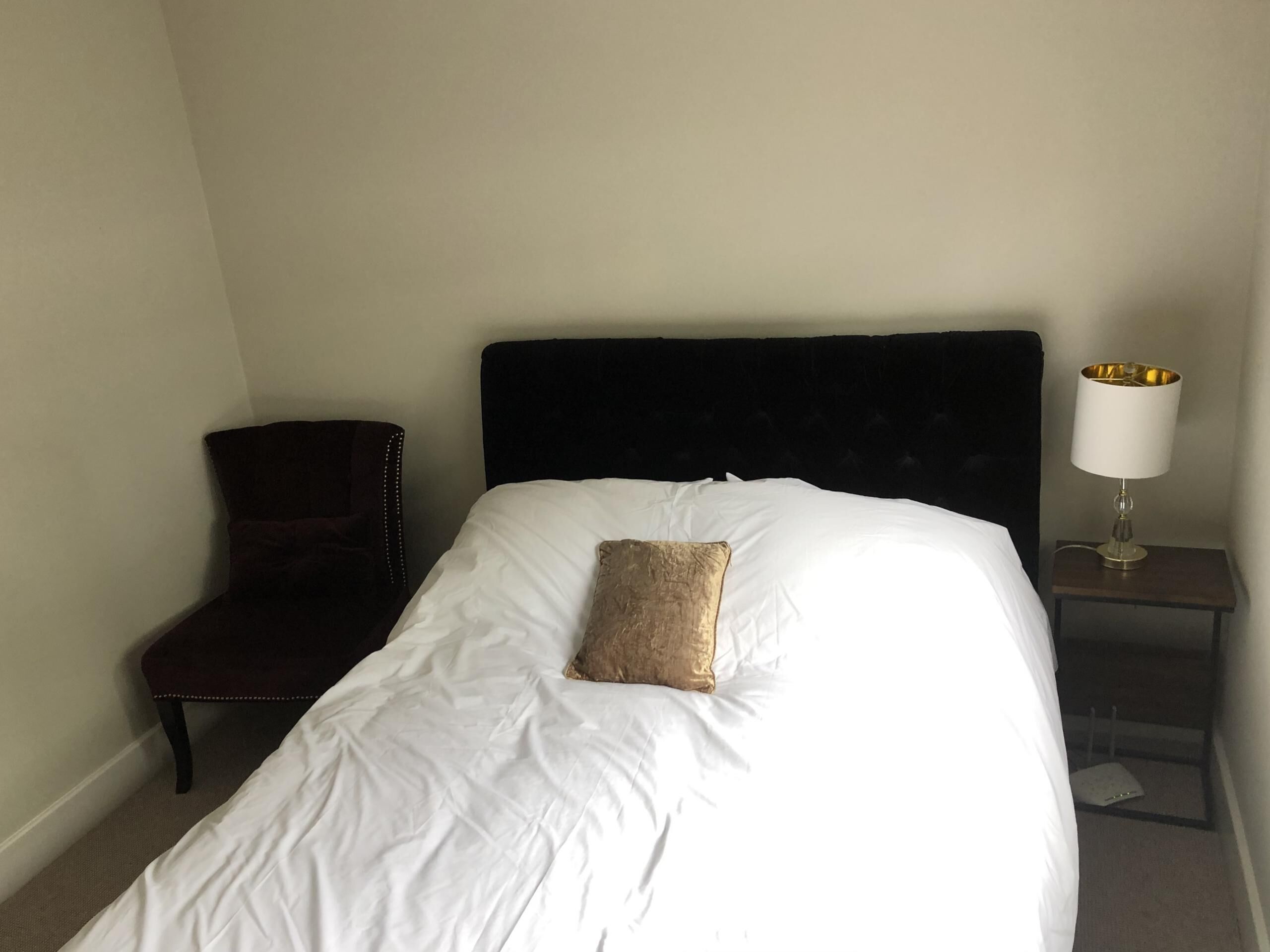 Ein ordentlich bezogenes Bett mit weißen Leinen und einem einzelnen Zierkissen, ein dunkel bezogener Sessel sowie ein Nachttisch mit einer Lampe.