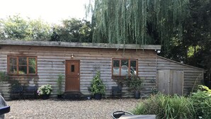 A rustic wooden cabin with a gravel driveway, a wooden door, and windows. There are two potted plants and a bench outside.