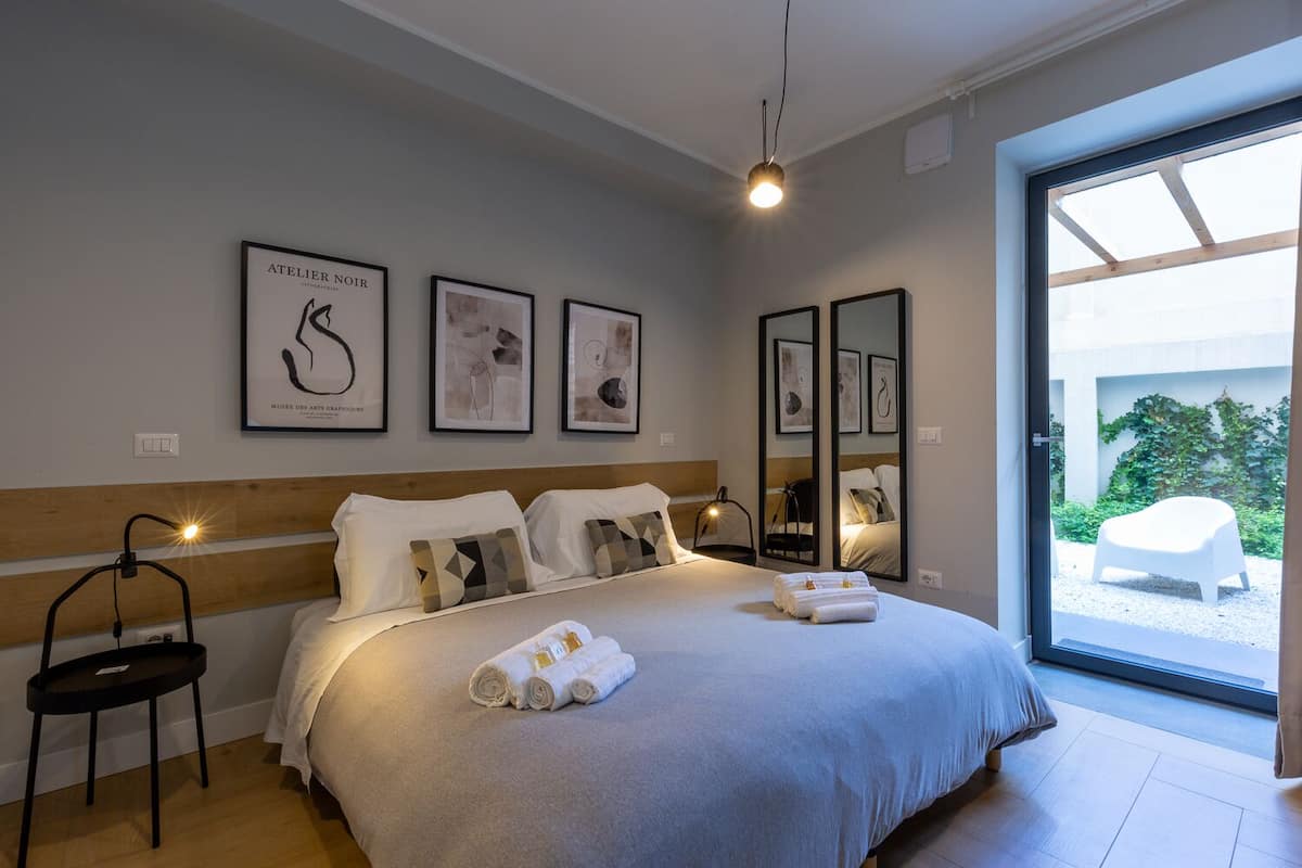 A modern hotel room with a large bed, a bedside table, a chair, and artwork on the walls.