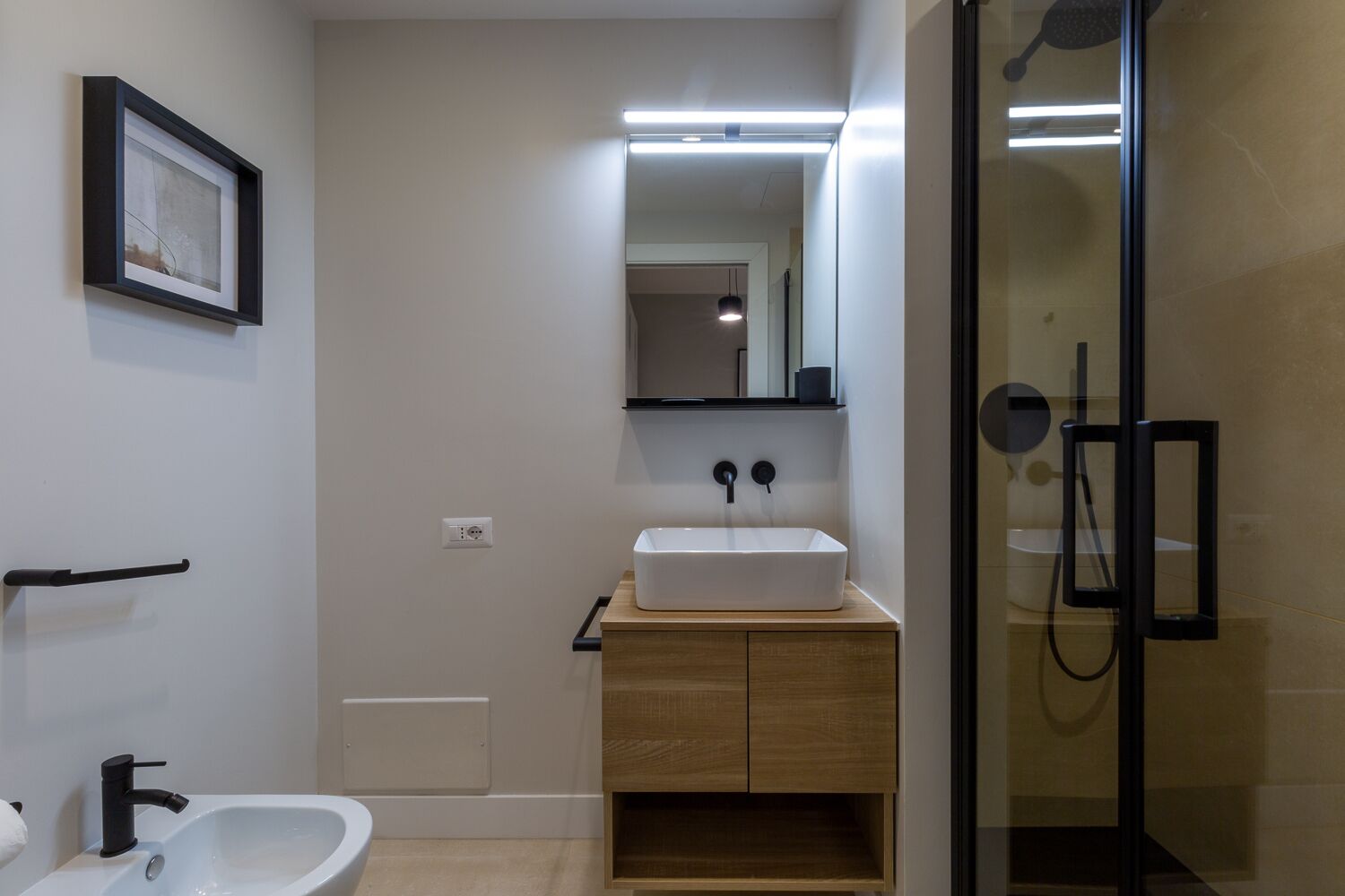 Ein modernes Badezimmer mit einem weißen Waschbecken, einem Holz-Badmöbel und einer Duschkabine aus Glas. An der Wand hängt ein gerahmtes Bild.