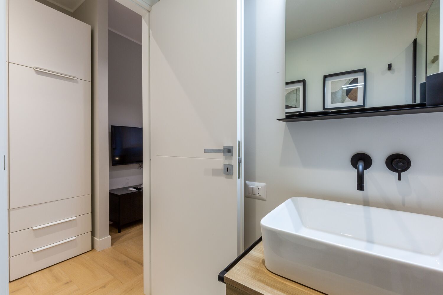 Une salle de bain moderne avec un lavabo blanc, un robinet noir et un miroir au-dessus. Une porte donne accès à une autre pièce.