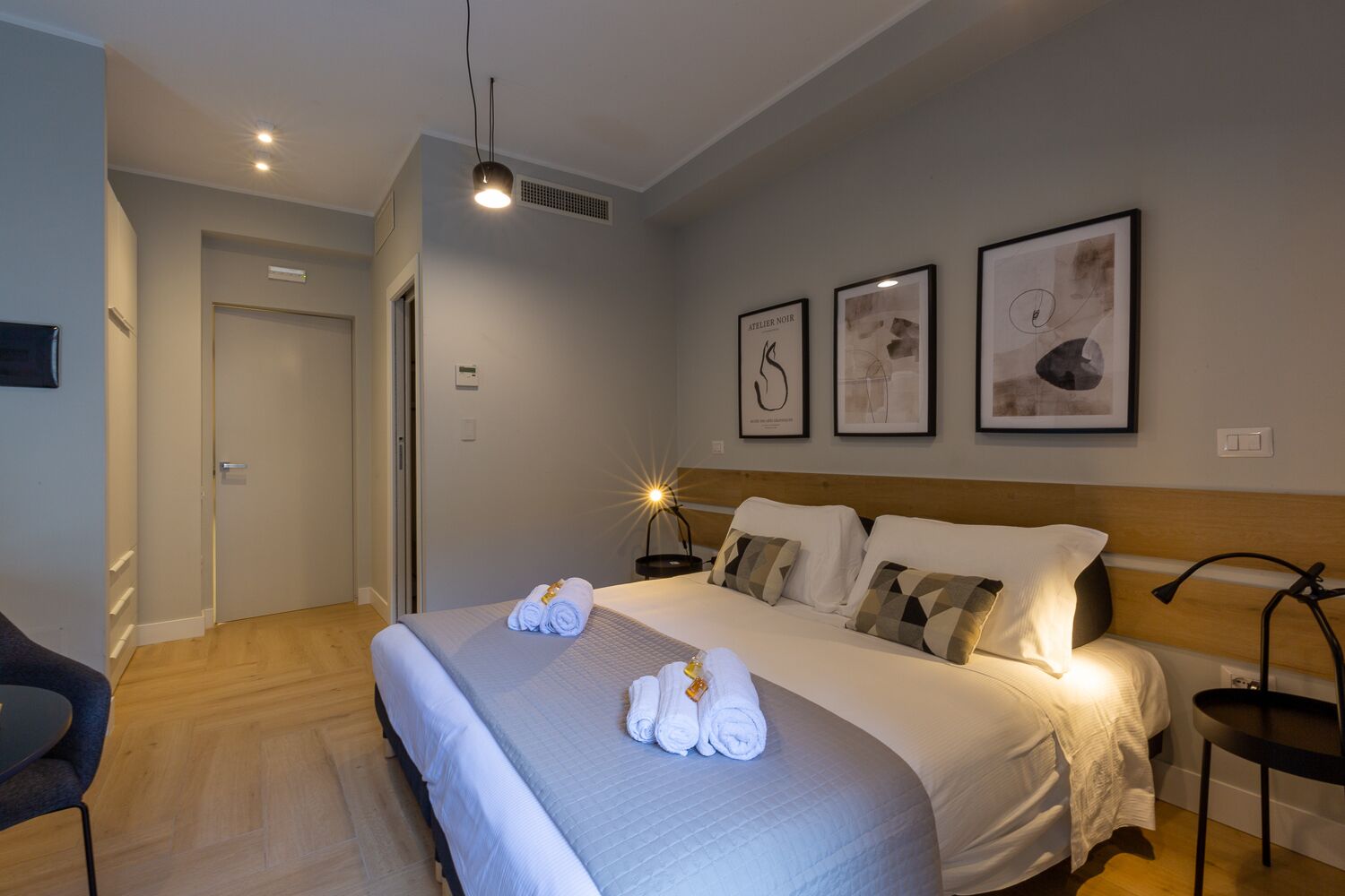 A hotel room with a large bed, a desk, and a chair. There are framed pictures on the wall and a ceiling light.