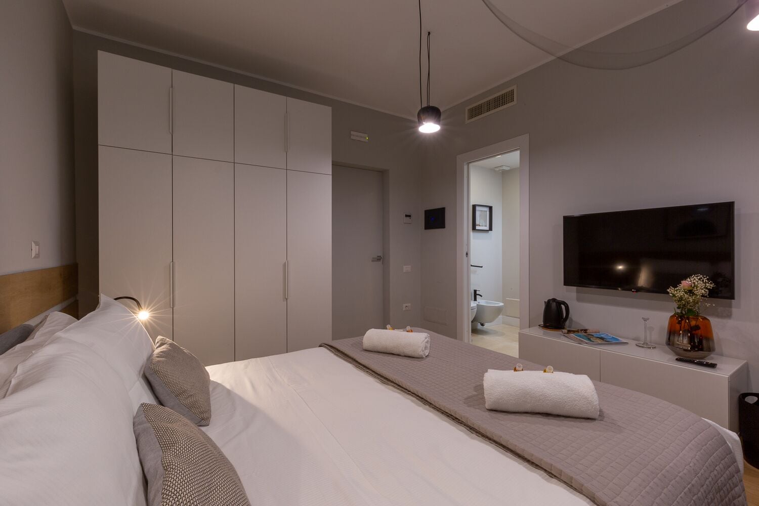 Una habitación de hotel moderna con una cama grande, un armario, un televisor y un baño visible a través de una puerta abierta.