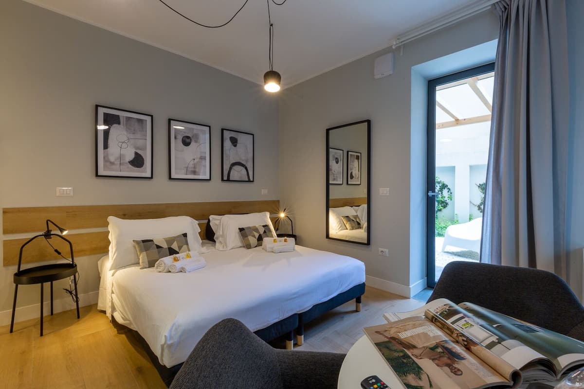 A hotel room with a bed, a chair, a table with magazines, and a window with curtains.