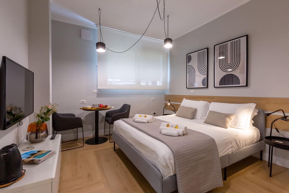 A modern hotel room with a large bed, a desk with a chair, a small table with a chair, and artwork on the walls.