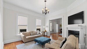 A spacious living room with a beige sofa, a blue ottoman, and a flat-screen TV mounted above a fireplace. - Spacious Midtown Home, Walk to Mardi Gras, Pets Ok
 (Mobile)