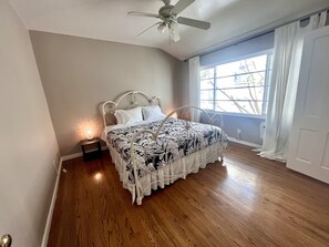Un dormitorio con cama, ventilador de techo, ventana con cortinas y mesita de noche con lámpara.