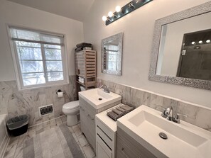 Un baño moderno con dos lavamanos, inodoro, tocador con cajones, espejo y ventana con persianas.