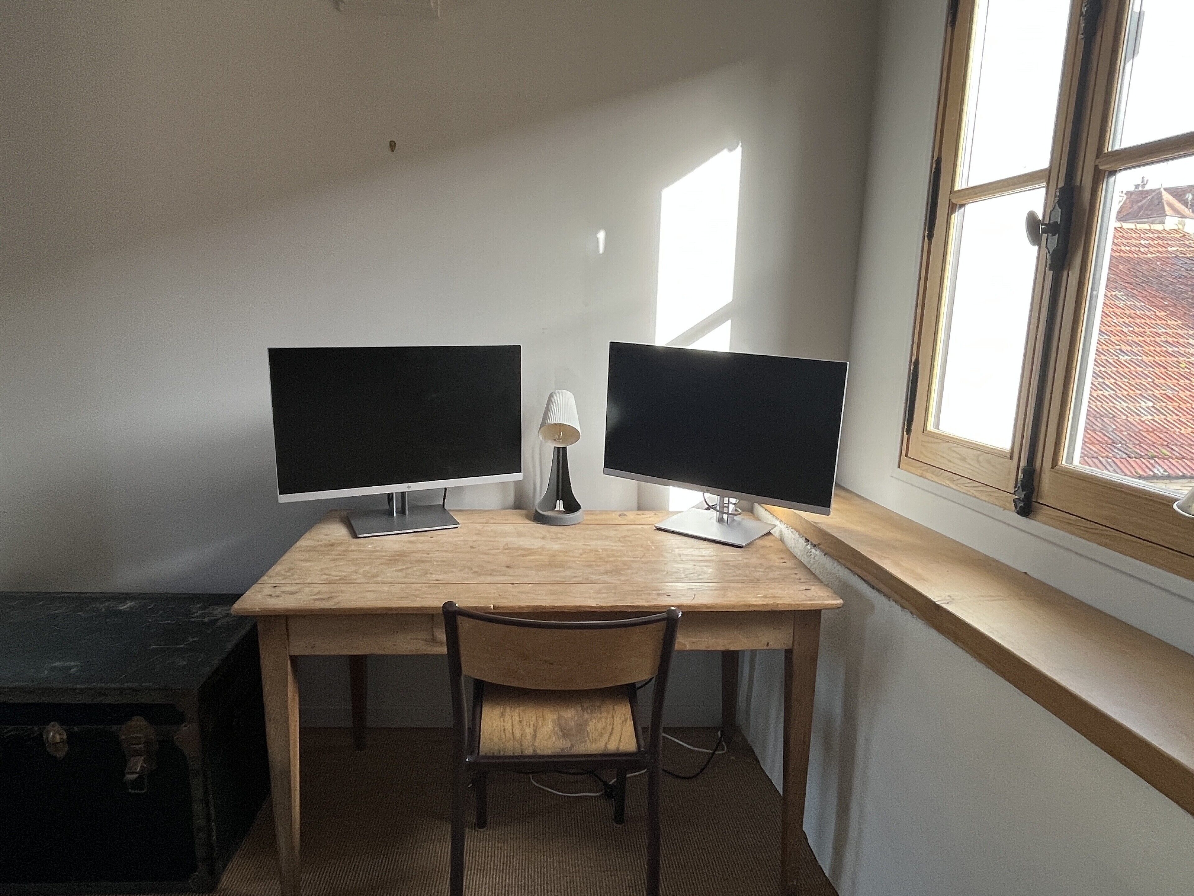 Ein Arbeitsplatz mit zwei Bildschirmen, einer Lampe und einem Stuhl an einem Holztisch neben einem Fenster.