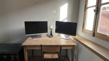 Ein Arbeitsplatz mit zwei Bildschirmen, einer Lampe und einem Stuhl an einem Holztisch neben einem Fenster.