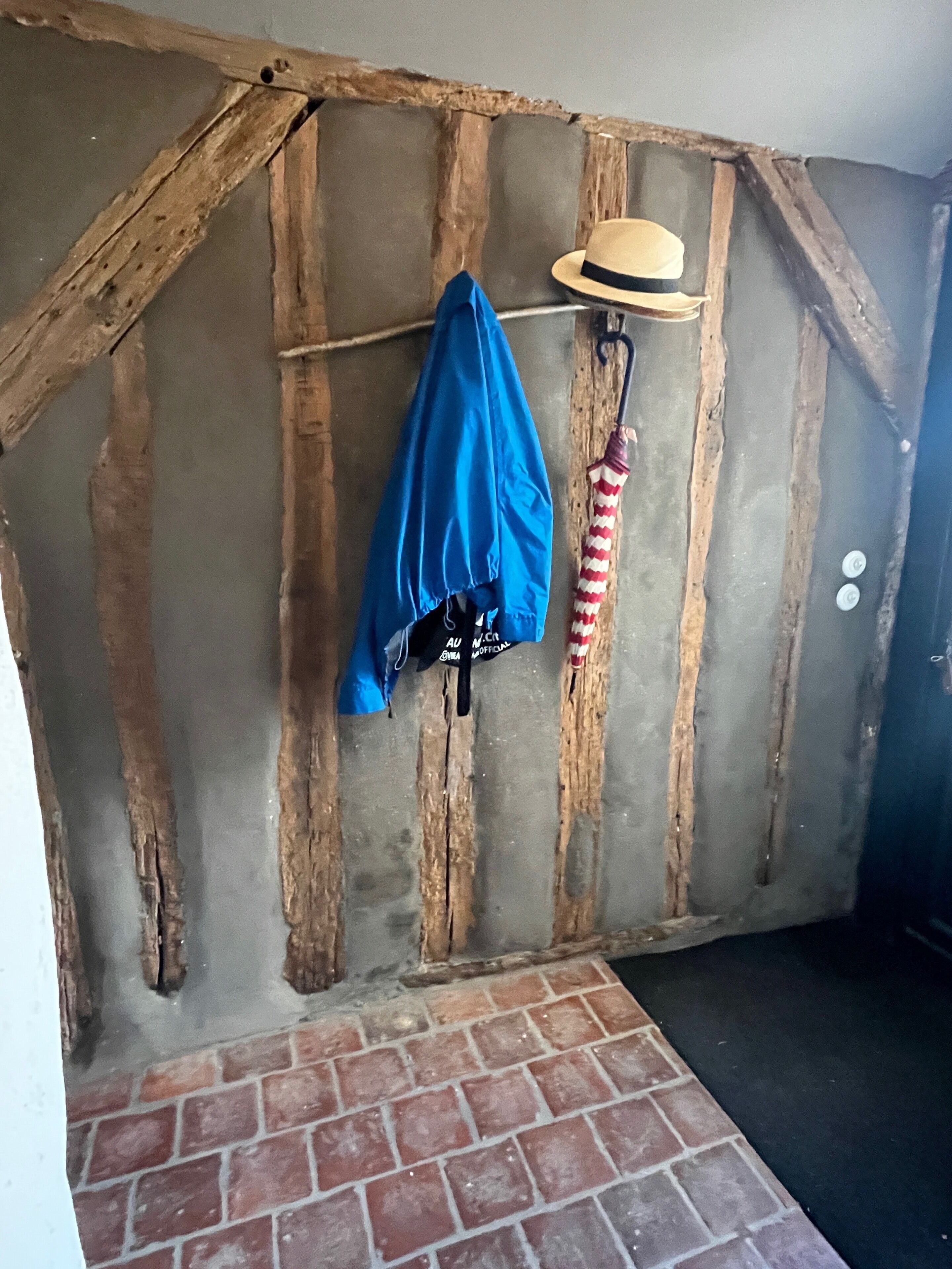 Una habitación rústica con vigas de madera a la vista, suelo de ladrillo, una chaqueta azul colgada de un perchero y un sombrero de paja en un soporte de pared.