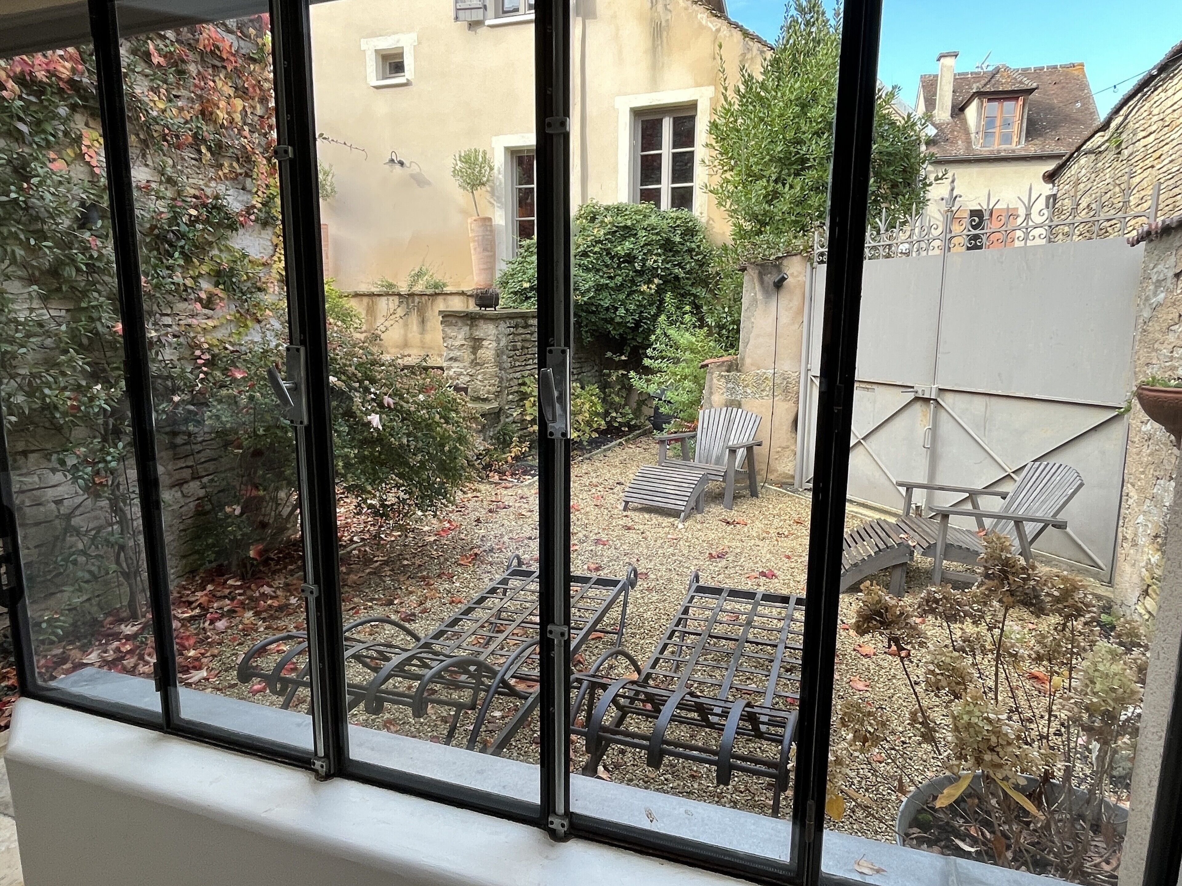 A view through a window of a courtyard with a metal grill, a wooden chair, and a stone wall.