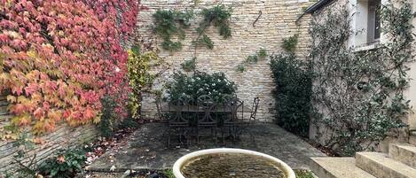 A stone building with a tiled roof, a garden with a birdbath, and a table and chairs set up outside.