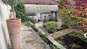 Un edificio de piedra con techo de tejas rojas, un jardín con diversas plantas y una maceta de terracota grande.